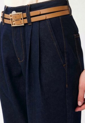 Pantalon en denim foncé avec plis et poches avant inclinées, porté avec une ceinture fine en cuir marron clair à double boucle, partie du torse et de la main d'une personne visible.