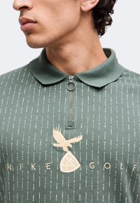 Νέος άνδρας που φοράει πράσινο πόλο Nike Golf με φερμουάρ και λογότυπο αετού, κοιτάζει αριστερά με εμφανή τον λαιμό και τη γνάθο.