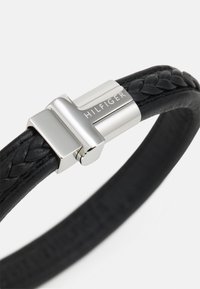 Pulseira de pele trançada preta com um fecho prateado gravado com "HILFIGER". Textura suave e design elegante e minimalista.