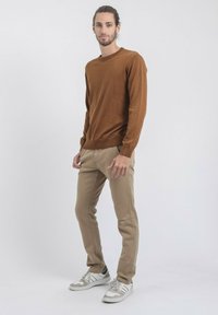 Pull long à manches longues marron, pantalon slim beige et baskets blanches avec des accents gris. Texture lisse et design décontracté. Fond neutre.