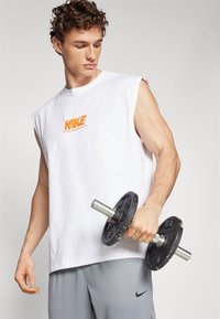 Witte mouwloze Nike-shirt met oranje logo; grijze sportshorts; houdt een zwart gewicht met zilveren handvat vast; neutrale achtergrond.