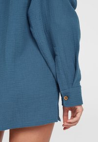 Blauwe, gestructureerde shirt met een ontspannen pasvorm. Heeft een knoopsluiting bij de manchetten en zijopeningen voor extra detail. Het materiaal lijkt lichtgewicht en ademend.
