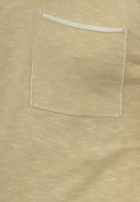 Beigen värinen kangas, jossa on teksturoitu pinta ja suorakaiteen muotoinen tasku, jonka reunoja korostaa valkoinen kontrastireunus.