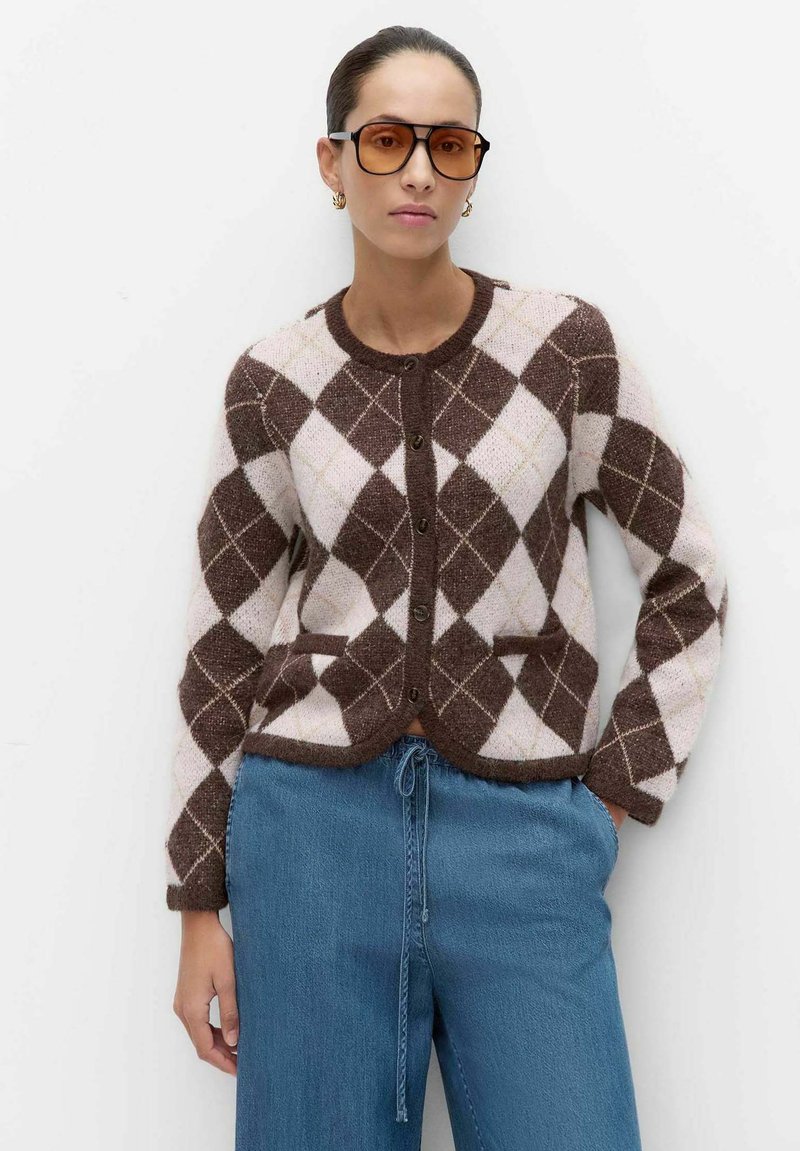 Kvinde iført brun og beige argyle cardigans, blå bukser med snøre, og store solbriller med ravfarvet glas, stående mod en ensfarvet hvid baggrund.