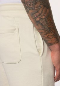 Joggers couleur crème en tissu doux, dotés de poches latérales et d'un logo en relief subtil près de la taille. Design texturé visible.