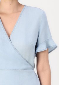 Robe portefeuille bleu clair à manches courtes volantées portée par une personne, montrant le haut du torse et le décolleté sur un fond uni.