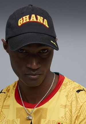 Joven con gorra negra con "GHANA" en amarillo, camiseta deportiva amarilla con estampado y ribete rojo, y un collar de cadena plateada con dijes.