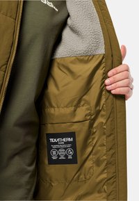 Olivgrön vadderad jacka med grå fleecefoder, med en svart etikett som beskriver TEXATHERM Core-teknik.