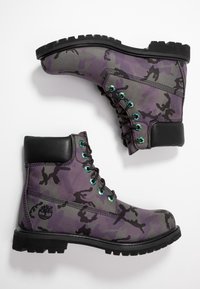 Botas con un diseño de camuflaje en púrpura y gris, con detalles en cuero negro, cordones y ojales verdes. Suelas de goma resistentes para mayor tracción.