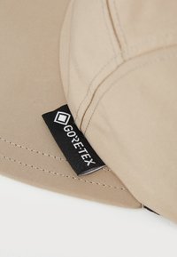 Beige színű, látható varratokkal készült anyagú sapka, melynek oldalán egy fekete GORE-TEX címke van belevarrva.