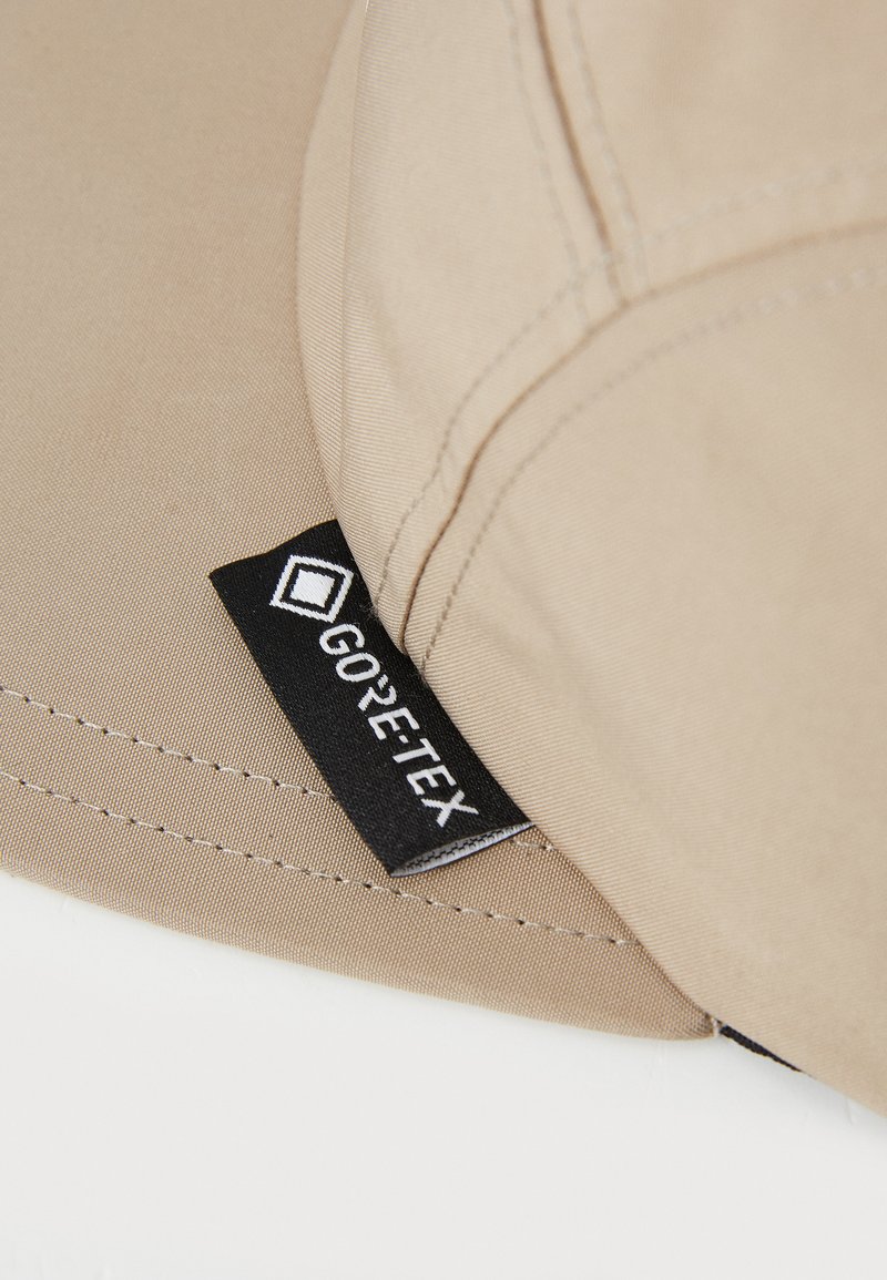 Beige pet gemaakt van stof met zichtbare stiksels, voorzien van een zwart GORE-TEX-label dat aan de zijkant is genaaid.