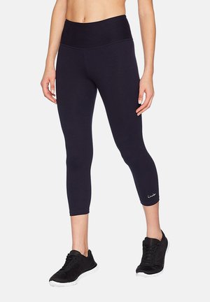 Black capri leggings made of stretchy fabric with a high waistband and subtle logo detail near the hem, paired with black athletic shoes.
