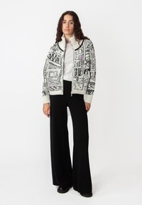 Gebreide cardigan in crème, met zwarte en lavendelkleurige geometrische patronen, hoge kraag, volledige rits en geribde manchetten. Gepaarde met zwarte wijdere broeken.