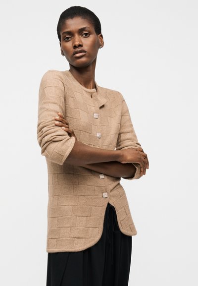 Vrouw met kort haar staat met gekruiste armen, draagt een beige gebreid vest met vierkante knopen en zwarte broek.