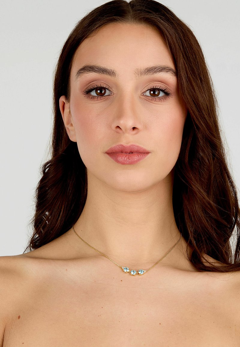 Collana in oro con tre gemme blu, superficie liscia, chiusura a moschettone e design delicato che si appoggia su una clavicola nuda.