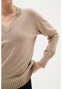 Beige stickad tröja med V-ringning, ribbade manschetter och nederkant, med en mjuk textur. Modellen poserar med en hand på höften.