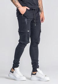 Joggers de carga negros con un corte ajustado, que cuentan con bolsillos laterales, detalles de tela desgastada y una cinturilla con cordón, combinados con zapatillas blancas.