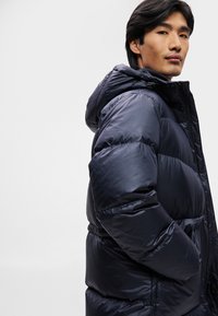Casaco puffer azul-marinho com capuz, tecido brilhante, design acolchoado, bainha elástica e bolsos laterais; robusto e quente para o tempo frio.