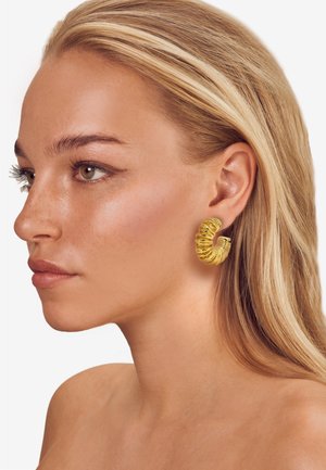 Goldfarbene Ohrringe in Form von Creolen mit einem gedrehten Design. Glatte, glänzende Oberfläche mit Rillen, die einen strukturierten Look erzeugen. Leicht anmutendes Erscheinungsbild.