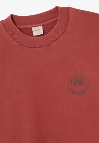 Πορτοκαλί φούτερ με στρογγυλή λαιμόκοψη και ριμπ τύπου κολάρο, με ένα μικρό σκούρο λογότυπο ενός δέντρου και κείμενο στην περιοχή του αριστερού στήθους.