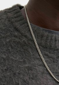 Tæt på mørkhudet person iført en struktureret mørkegrå sweater og en sølv kædder halskæde over en grå skjortekrave.