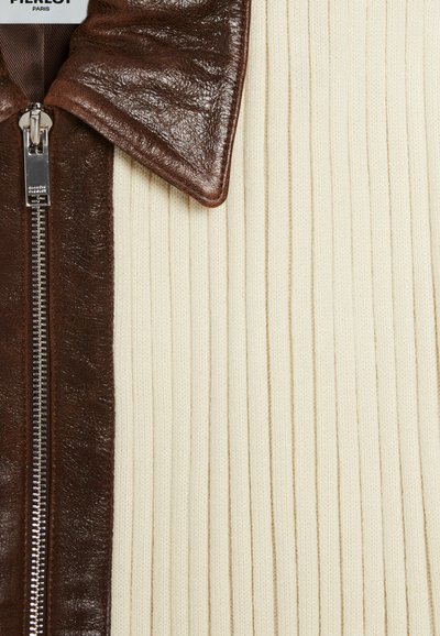 Hnedý kožený golier so zipsom cez krémovú vertikálne pruhovanú látku. Textúrovaný povrch a viditeľné švy. Značková etiketa na golieri.