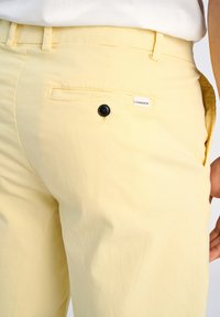 Pantalons jaune clair avec deux poches arrière, l'une comportant un bouton noir et une étiquette de marque. Texture de tissu douce et coupe ajustée.