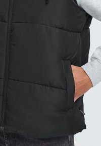 Schwarze gepolsterte Weste mit Reißverschluss, seitlichen Taschen und einem strukturierten Stoff. Darunter befindet sich ein hellgraues Langarmoberteil.