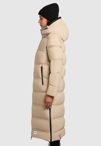 Ilgas smėlio spalvos pūkinis paltas su kapišonu, juodais užtrauktukų akcentais ir siuvinėtu tekstūru. Derinamas su juoda megzta kepure.