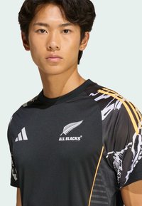 Černý sportovní dres s bílými a oranžovými akcenty, logem listu a strukturovaným designem na rukávech. Vyrobeno z lehkého, prodyšného materiálu.