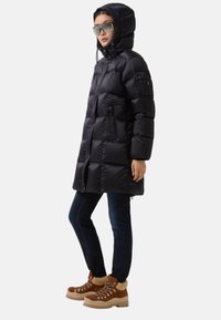 Zwarte pufferjas met een capuchon, voorzien van een gestikt ontwerp, ritszakken, gecombineerd met donkere jeans en bruine veterschoenen.