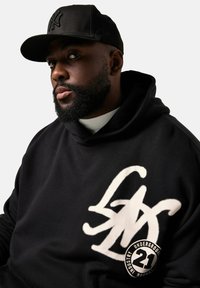 Sudadera negra con un gran diseño de logo blanco en el frente, combinada con una gorra de béisbol negra. El tejido parece suave y tiene un corte relajado.
