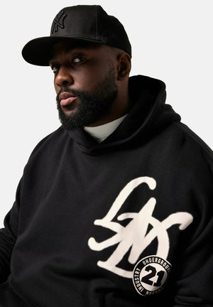 Schwarzer Hoodie mit einem großen weißen Logo-Design auf der Vorderseite, kombiniert mit einer schwarzen Baseballkappe. Der Stoff scheint weich zu sein und hat eine lockere Passform.