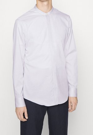 Mann trägt ein helles lavendelfarbenes Langarmhemd mit Mandarinkragen und verdeckten Knöpfen sowie dunkle Hose vor einem einfarbigen Hintergrund.