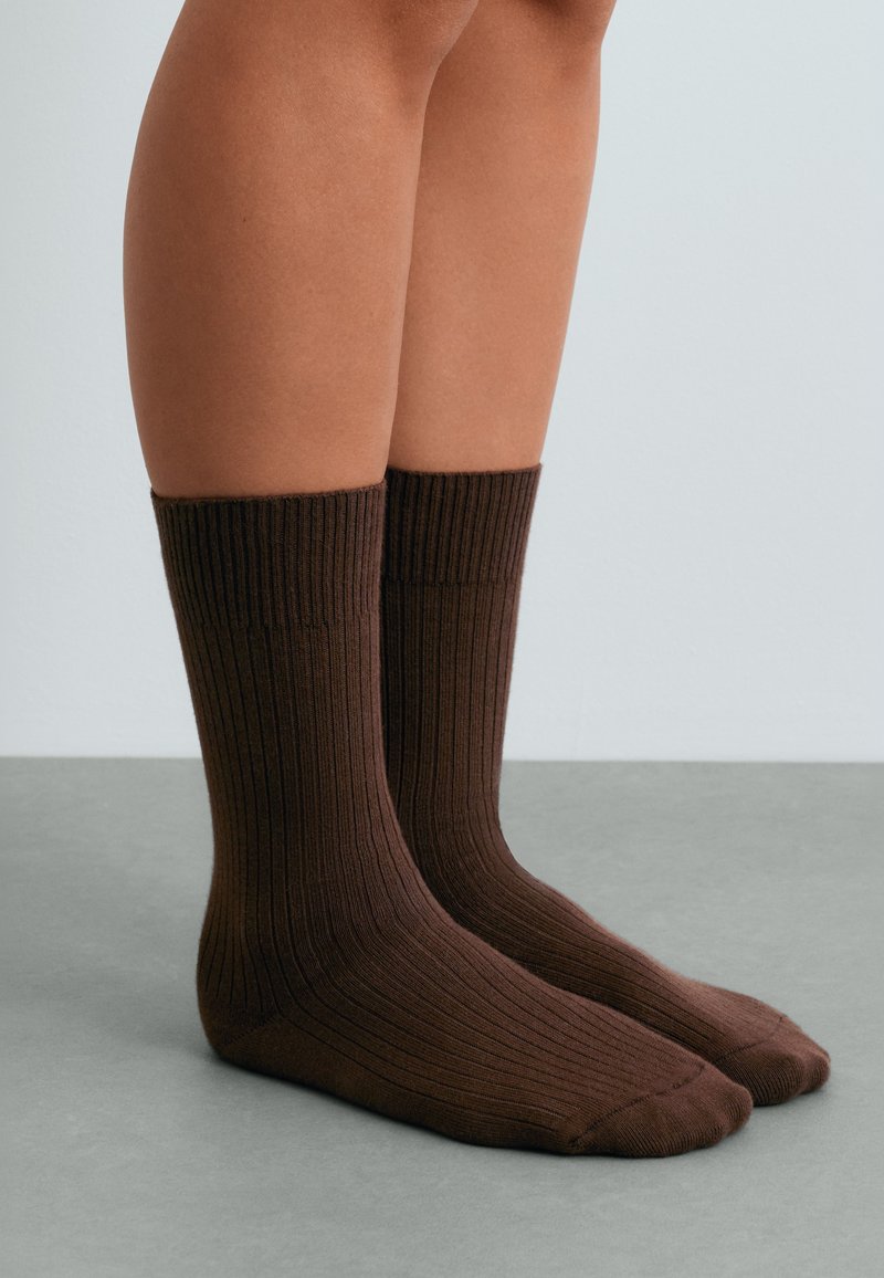 Chaussettes brunes côtelées jusqu'au mollet portées sur des pieds posés sur une surface grise avec un fond clair.
