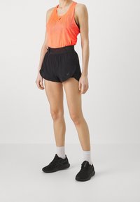 Helles orangees Mesh-Top mit geometrischem Muster, kombiniert mit schwarzen Sportshorts. Schwarze Turnschuhe und weiße Socken runden das Outfit ab.