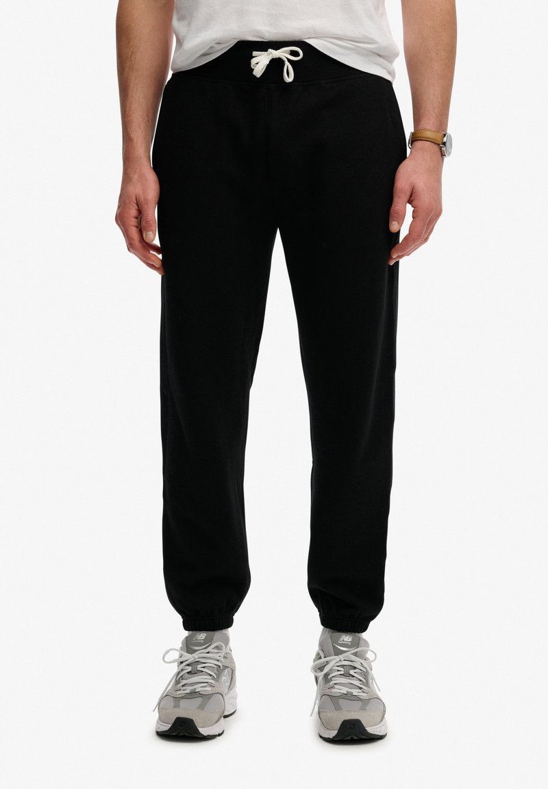 Mężczyzna w czarnych spodniach dresowych ze ściągaczem i szarych butach New Balance, stojący z rękami luźno opuszczonymi wzdłuż ciała, na białym tle.