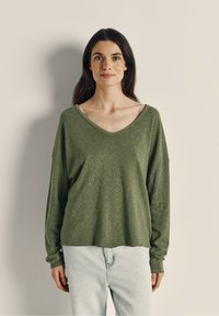 Olīvzaļš ilgu piedurkņu V-veida kakla tops, kas izgatavots no mīkstas, vieglas auduma. Tam ir brīvs piegriezums un pazeminātas plecu šuves. Saskaņots ar gaišām džinsām.