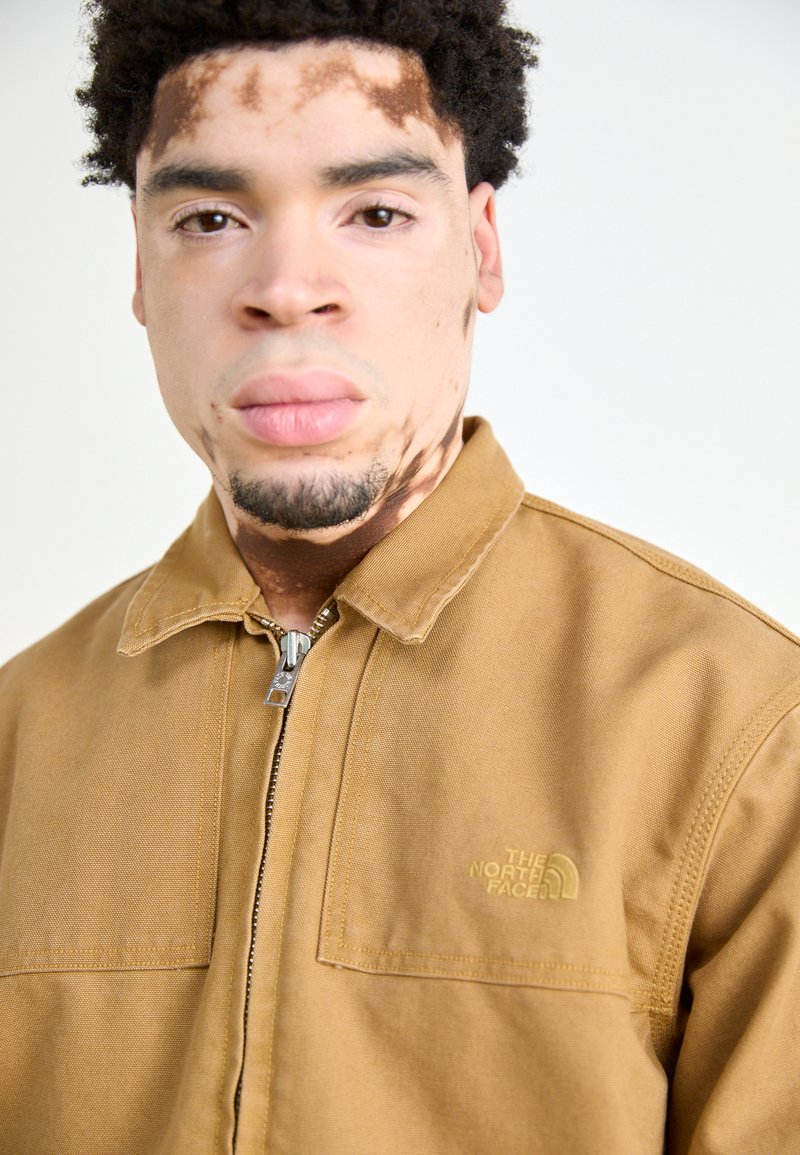 Chaqueta de lona marrón con cremallera, que cuenta con un cuello y dos bolsillos en el pecho. Pequeño logo amarillo en el lado izquierdo. Textura suave.