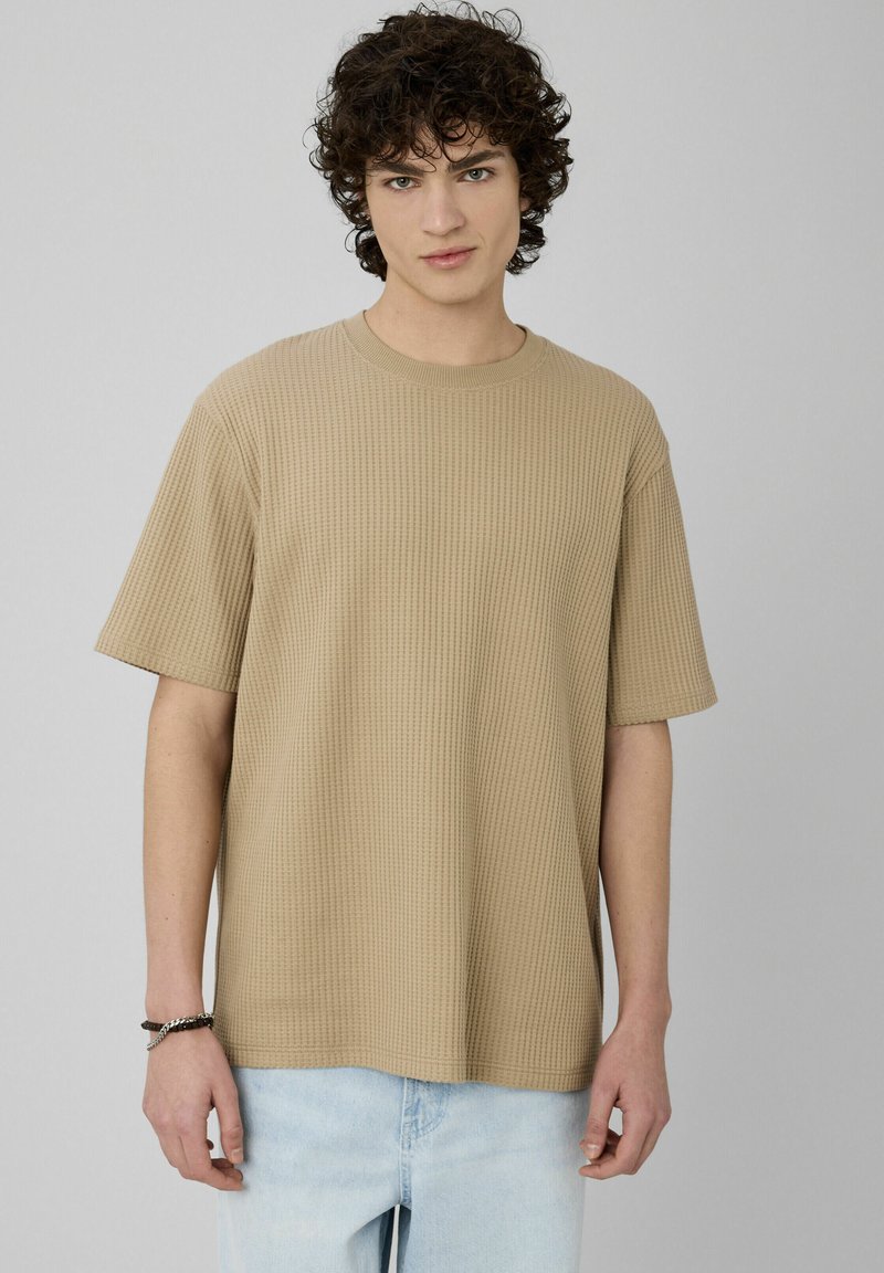 T-shirt beige à manches courtes côtelées avec un col rond. Texture lisse, coupe décontractée, associée à un jean bleu clair.