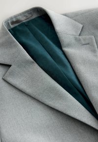 Blazer gris claro texturizado con forro interior de patrón verde oscuro, mostrado doblado enfocándose en las solapas y detalles del cuello.