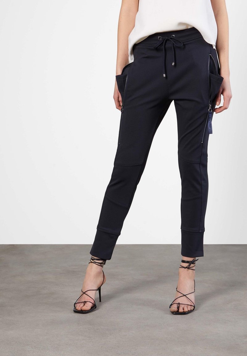 Jogging en navy en tissu extensible avec des zips sur les côtés, des poches avant et une taille ajustable par cordon, complété par des sandales à talons hauts noires avec des lanières.