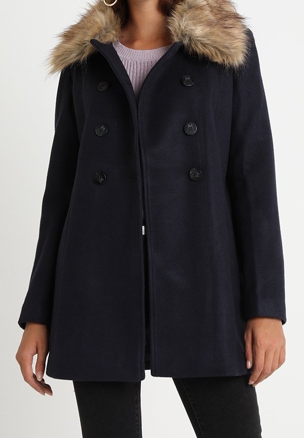 Femme portant un manteau noir croisé avec une capuche bordée de fourrure, par-dessus un pull violet clair et un pantalon noir.