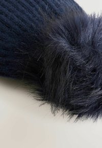 Niebieska czapka dziergana, ozdobiona dużym, miękkim pomponem z sztucznego futra w ciemniejszym odcieniu. Tekstura jest prążkowana, co podkreśla kontrast materiałów.
