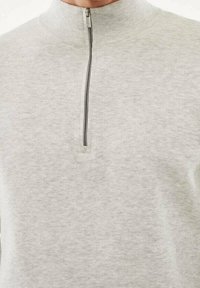 Maglione zip-up grigio chiaro realizzato in tessuto morbido. Presenta un colletto alto e una cerniera frontale con una linguetta in tonalità argento. Superficie semplice e testurizzata.