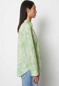 Camicia a maniche lunghe verde chiaro con motivo floreale, colletto button-down e spacchi laterali, abbinata a jeans blu chiaro.