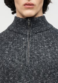 Gros plan sur une personne portant un pull en tricot gris foncé avec une demi-fermeture éclair et un col montant, montrant uniquement les lèvres et le cou.