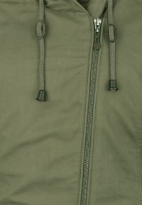 Olivengrøn jakke med lynlås foran, flettede snore og gummispidser. Jakken har en blød tekstur og ingen synlige mønstre eller hardware-detaljer.