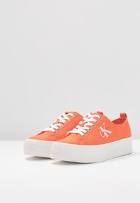 Orange canvas sneakers med vita gummisulor, med vita snören och en logotyp på sidan. Rundad tå och dragflik på sidan.