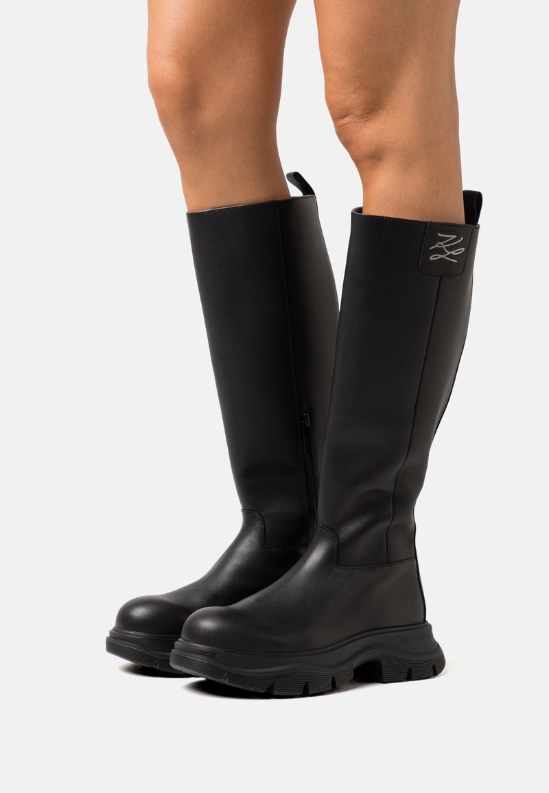 Jambes portant des bottes en cuir noir montant jusqu'aux genoux avec des semelles épaisses, des détails de couture, une fermeture éclair latérale et des languettes sur un fond clair.